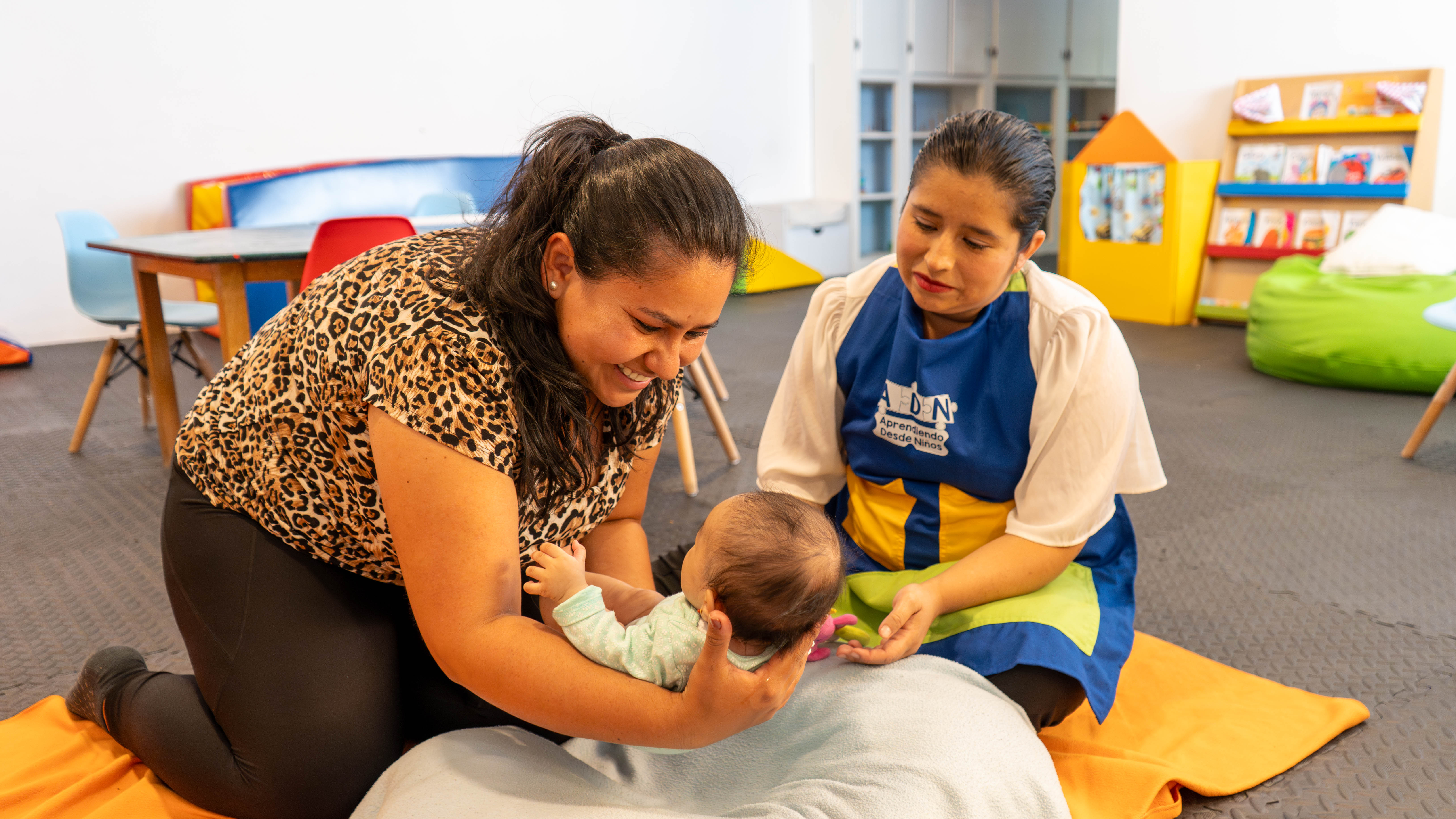 ADN program staff smiling while cradling a baby in the air above a beanbag sack on the floor