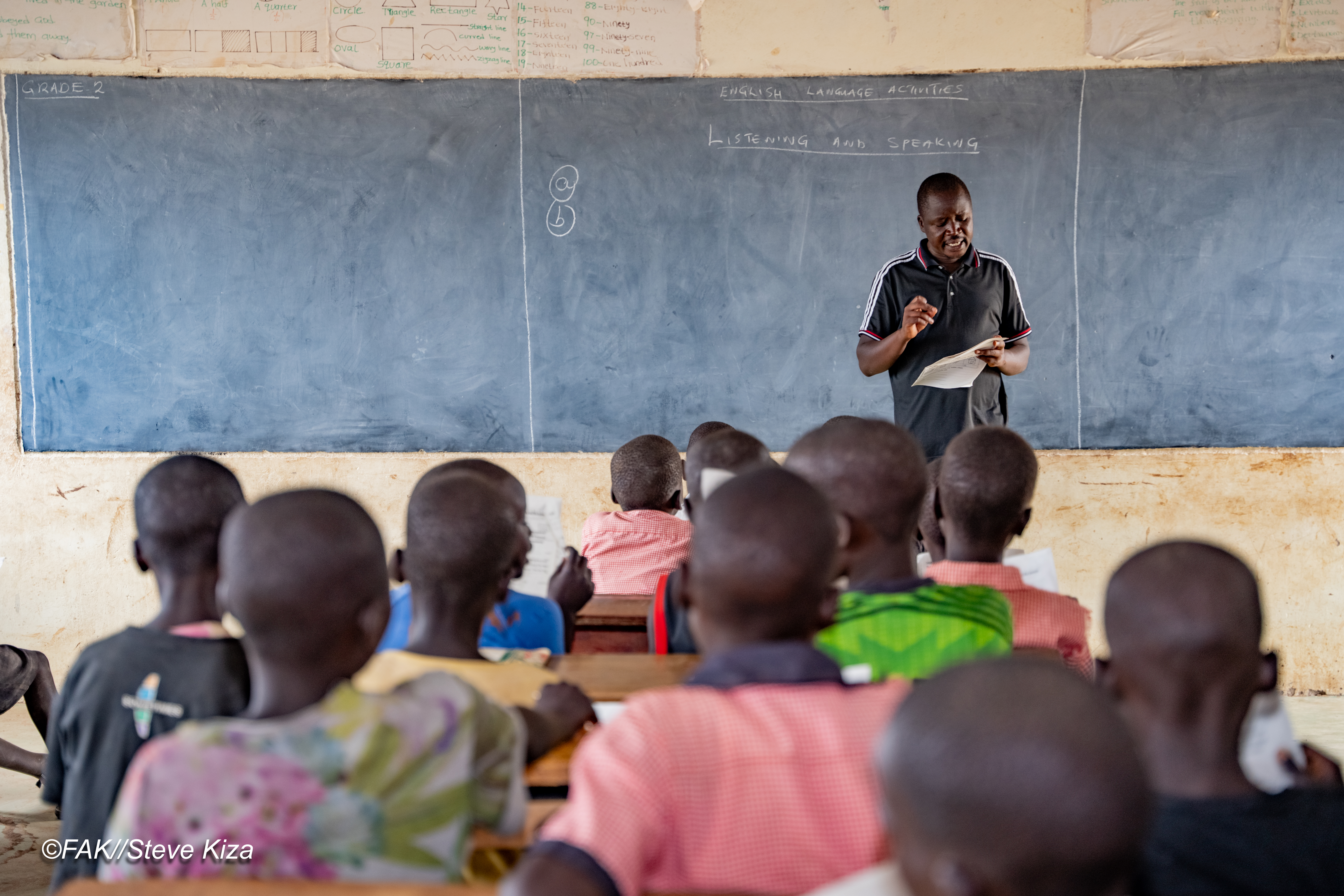 Photo of a teacher standing and speaking in front of a classroom full of students sitting at desks