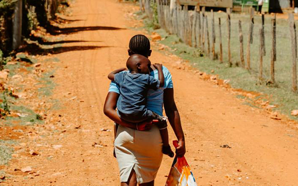 Photo d'une femme en Afrique marchant sur un chemin sablonneux, dos à l'objectif, portant un enfant sur son dos.