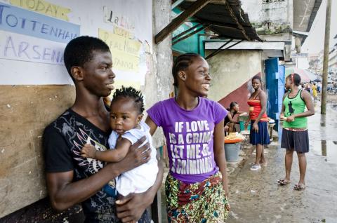 A couple of young people stand smiling with a child outside a building advertising soccer games in Monrovia, Liberia. © 2010 Glenna Gordon