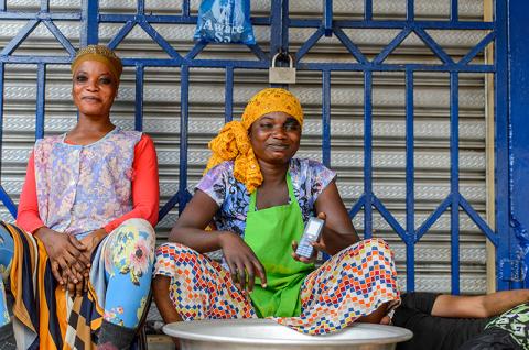 A Ghanaian woman in multicolor clothes and headscarf holds a cellphone at the Kumasi market in Ghana, while sitting next to another woman. © 2017 Anton Ivanov via Shutterstock