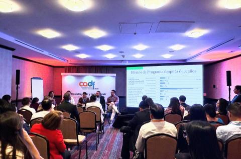 Presenters and audience during the panel session, with a projector screen showing a slide in Spanish being presented on the effects of the program after 3 years