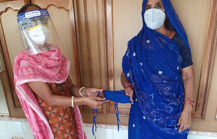 A woman in India wearing a face mask and face shield handing a cloth face mask to another woman also wearing a face mask